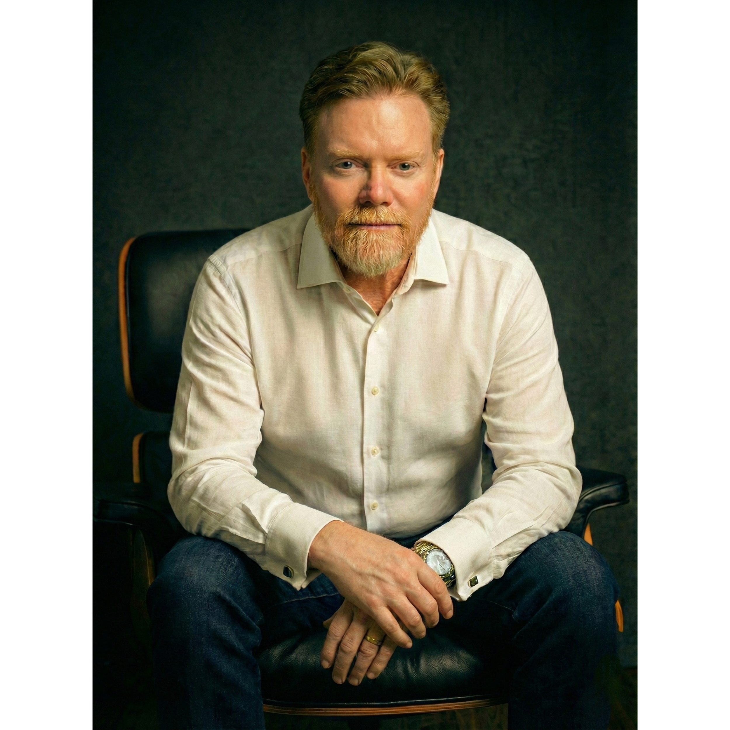 A man with red hair and a beard sitting on a black chair, wearing a cream-colored shirt and dark jeans in front of a dark textured background.