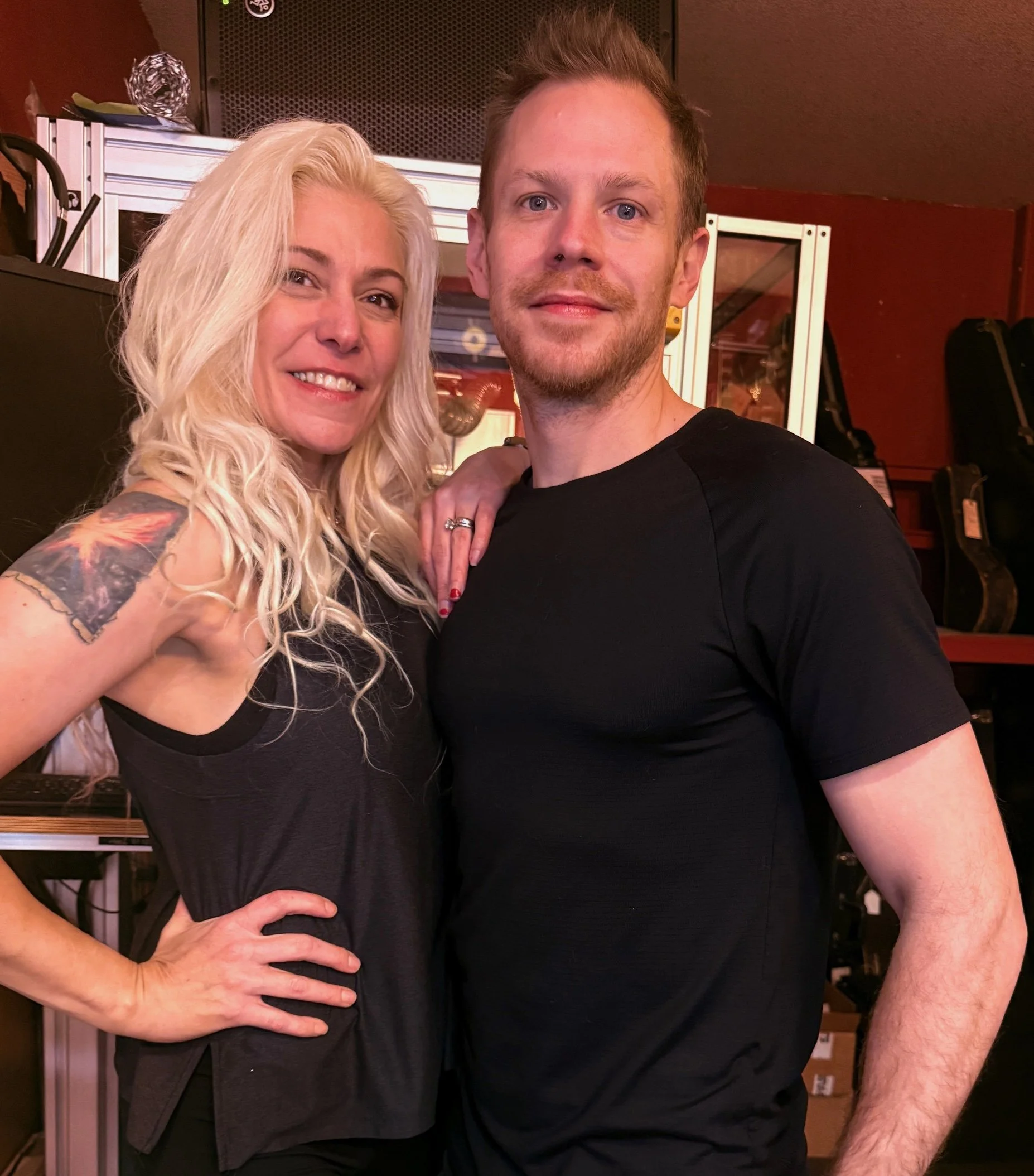 A smiling woman with long blonde wavy hair and a tattoo on her arm stands next to a man with short, light brown hair, both wearing black shirts, in a room with musical equipment and guitar cases in the background.