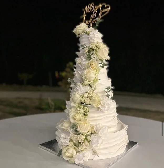 A tall wedding cake decorated with white flowers and a gold topper reading 'Happily Ever After.'