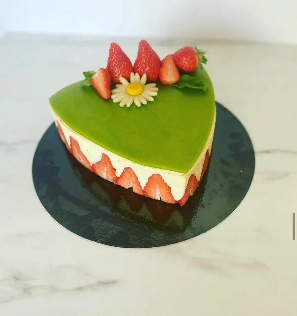 Heart-shaped cake with green top layer, strawberry slices around the side, topped with strawberries and a small white flower with a yellow center.