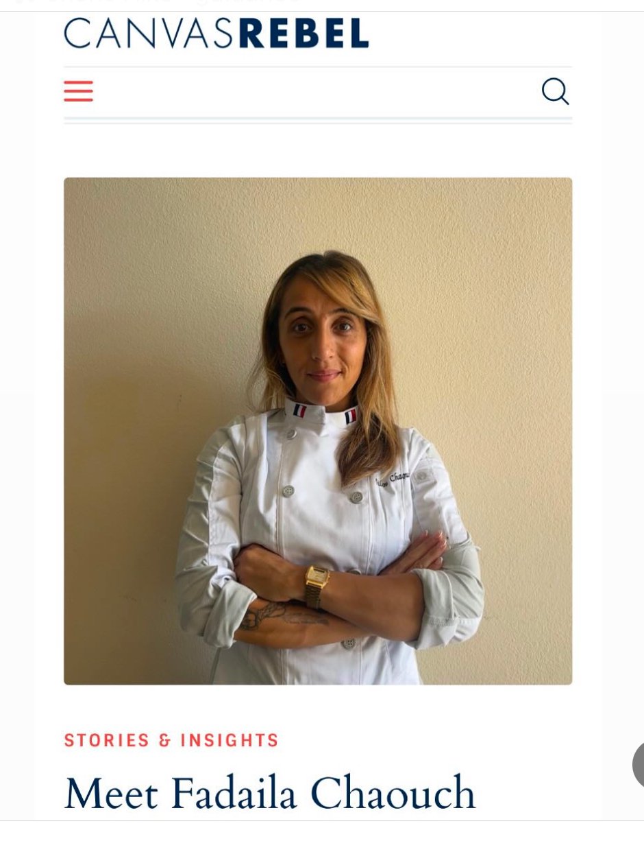 A woman with light brown hair wearing a white chef's coat with a French flag detail on the collar, standing against a beige wall with her arms crossed.