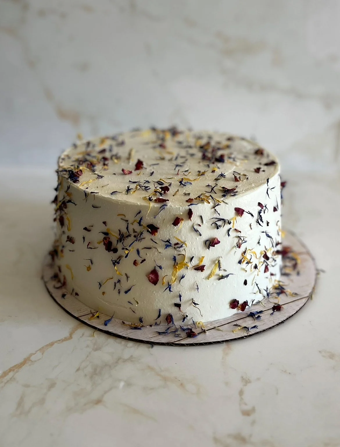 A round white frosted cake decorated with dried flower petals on top, placed on a white marble surface.