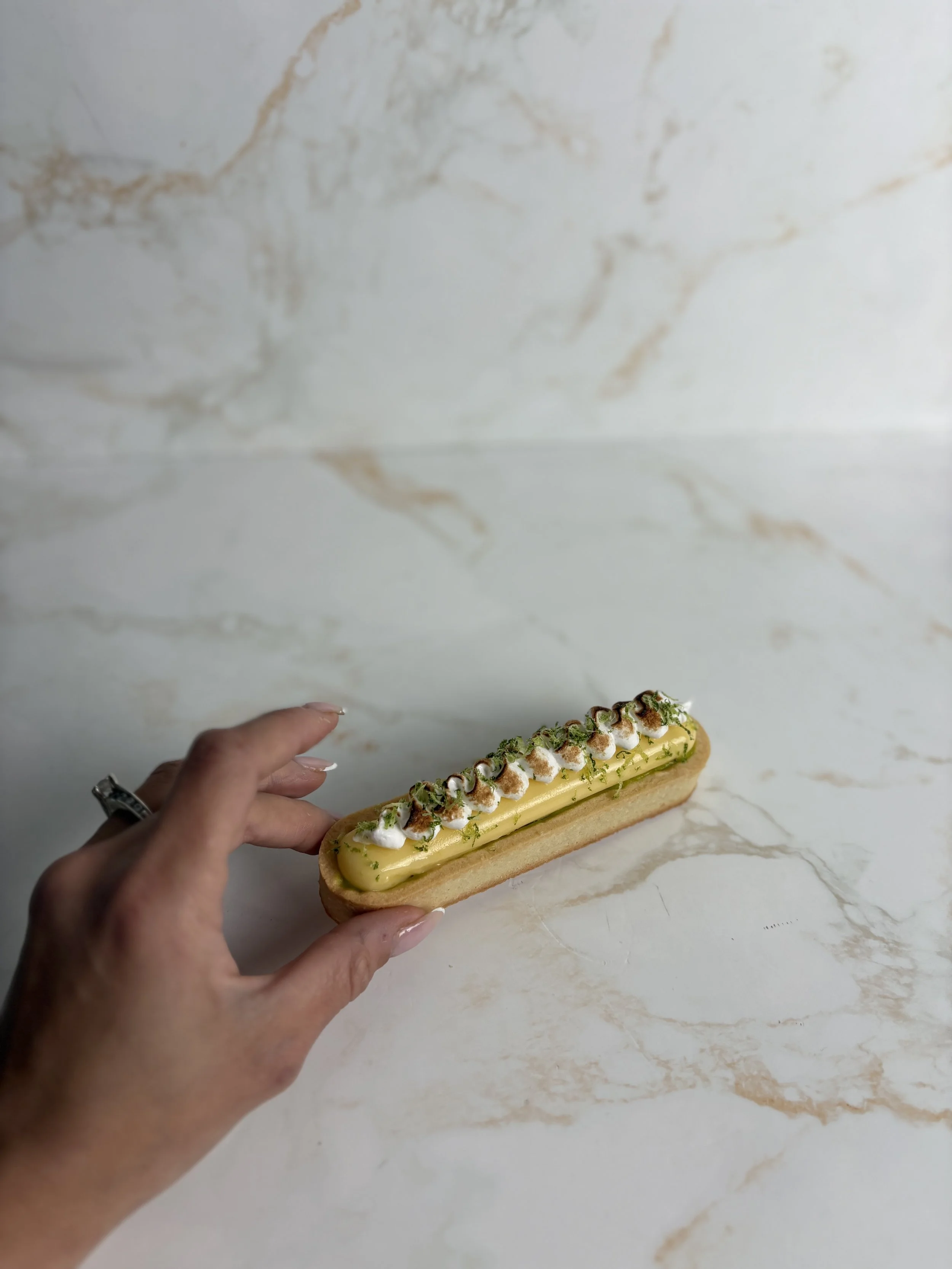 Person holding a long éclair pastry topped with yellow icing, whipped cream, chocolate, and green garnish on a light marble surface.