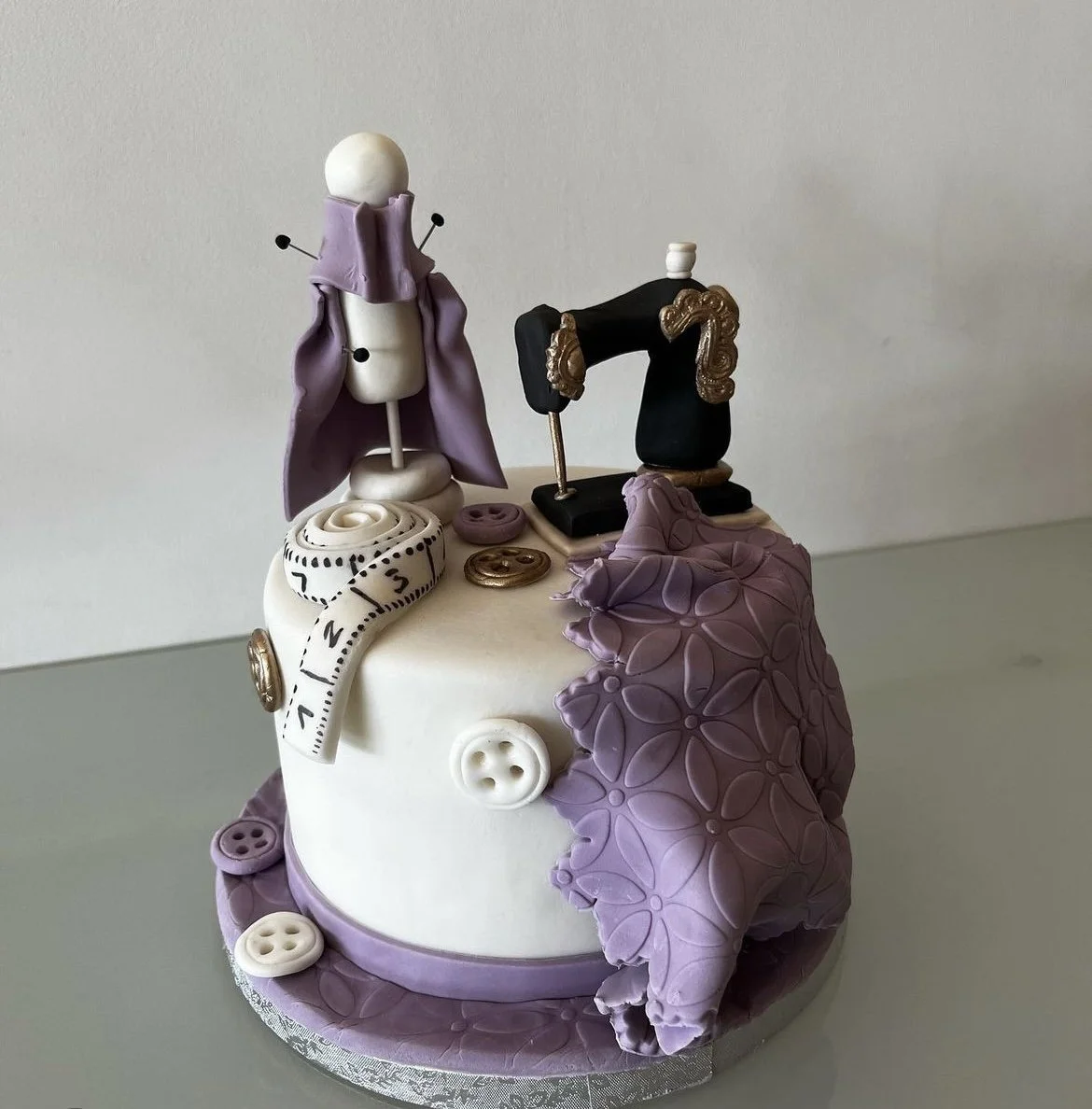 A decorated cake with sewing-themed decorations, including a mannequin dress form, a vintage sewing machine, buttons, a measuring tape, and purple floral accents.