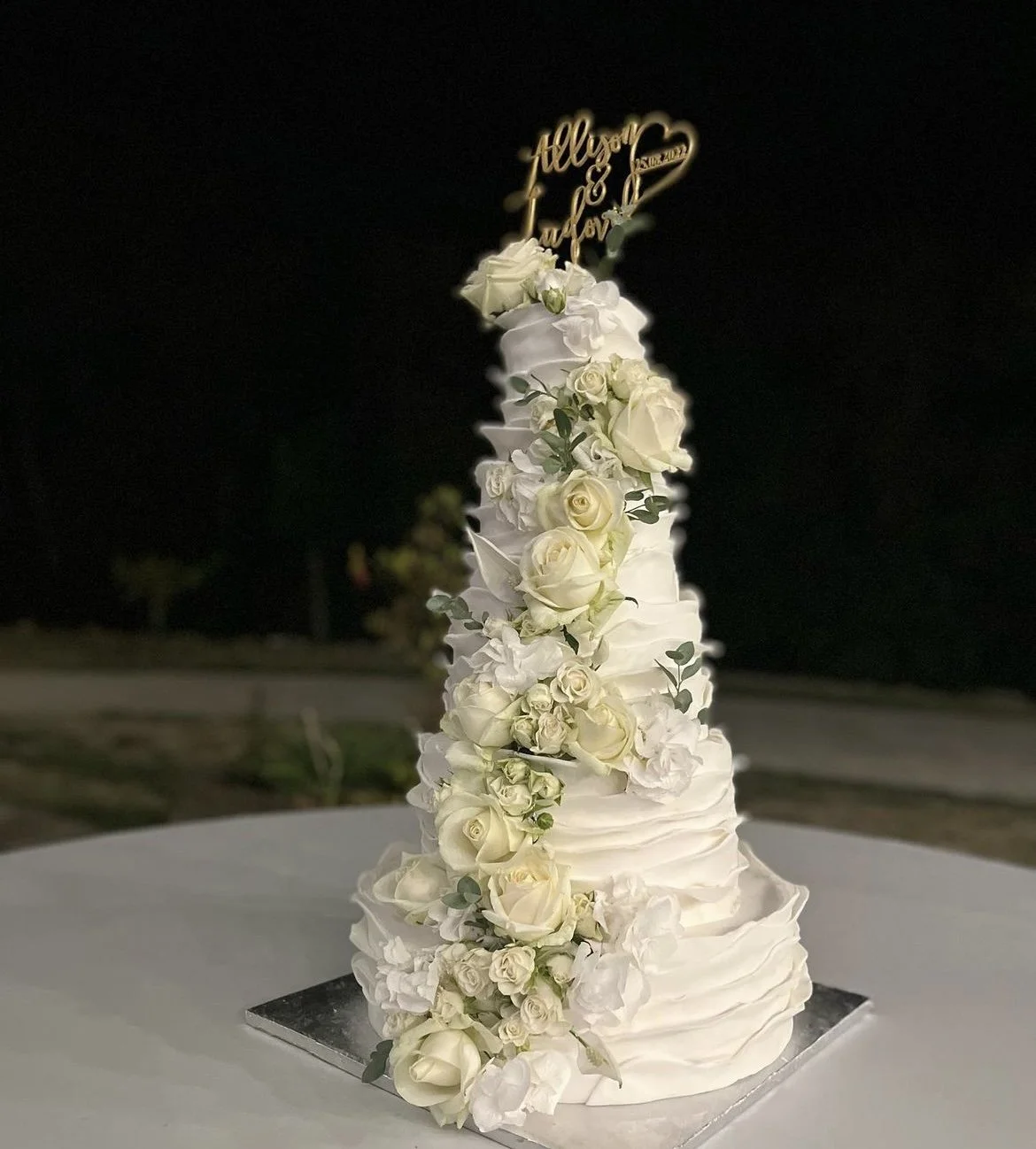 Tall wedding cake decorated with white roses and silver-green foliage, topped with a gold topper that says "Allison & J**" with a heart design, outdoors at night.