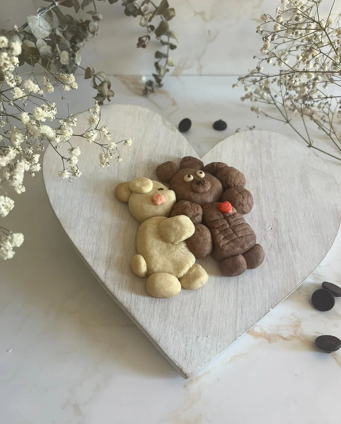 Decorative arrangement of animal-shaped cookies on a white wooden heart-shaped board, surrounded by dried flowers and scattered beans.