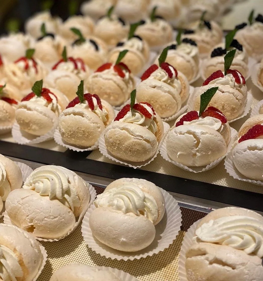 Close-up of white cream-filled sandwiches topped with red fruit and green leaf garnishes, arranged on a display tray.