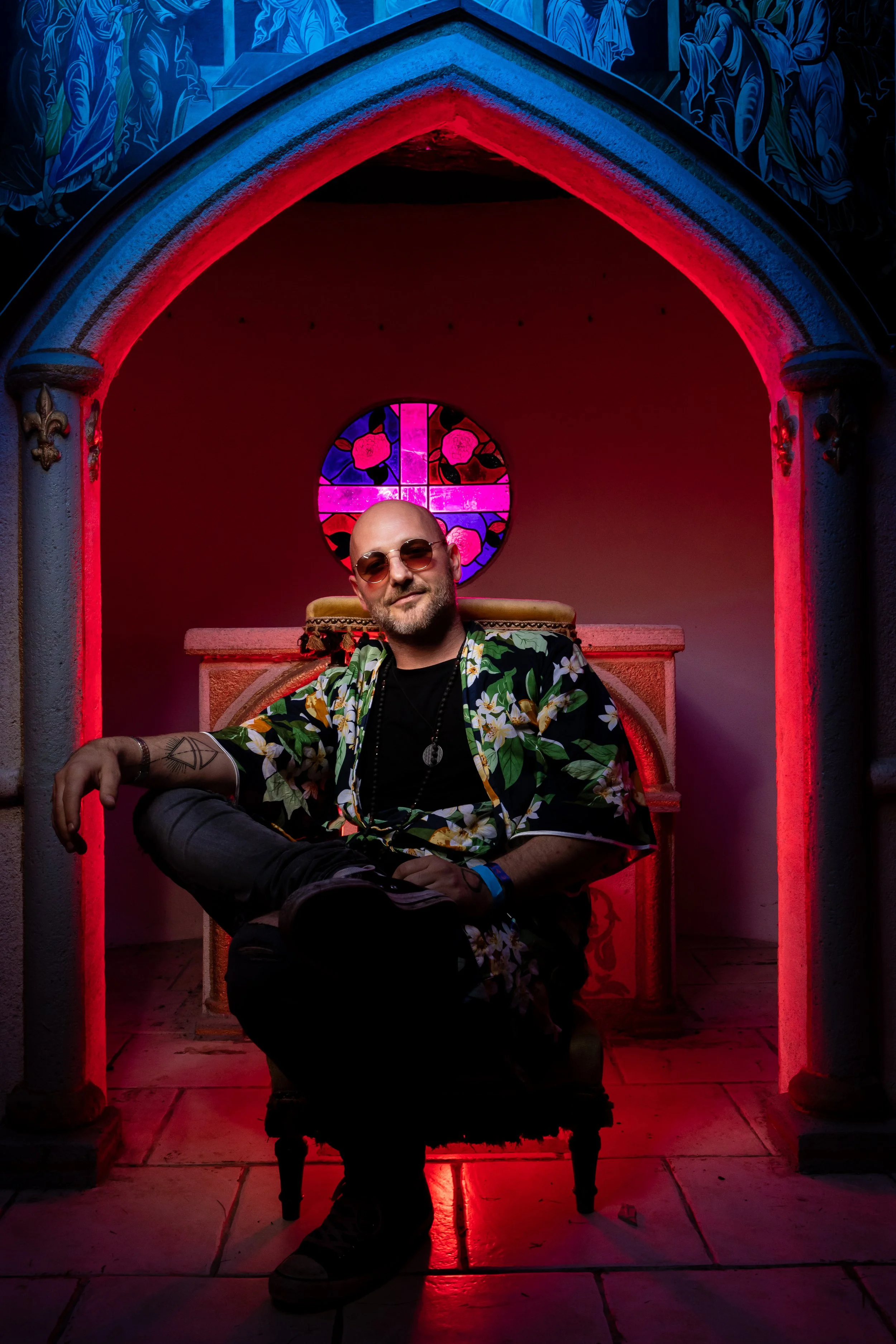 Glatzköpfiger Mann mit Sonnenbrille sitzt in einem Tempel mit bunten Fenstern, trägt einen hawaiischen Kimono, umgeben von rot-blauem Licht.