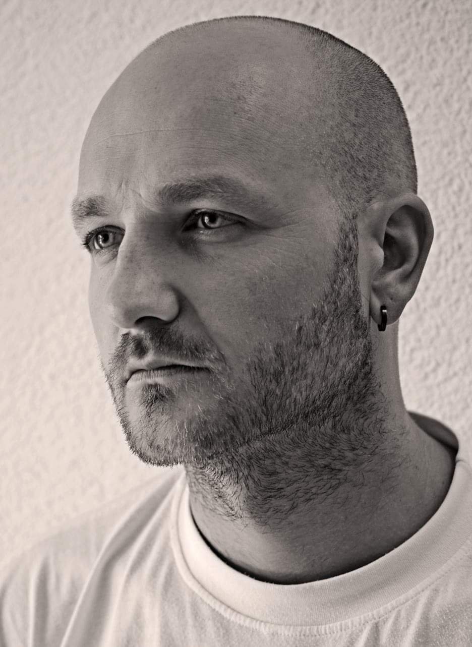 Mann mit blauen Augen und Glatze im weißem, reinem T-Shirt, hinter einer rauen Wand