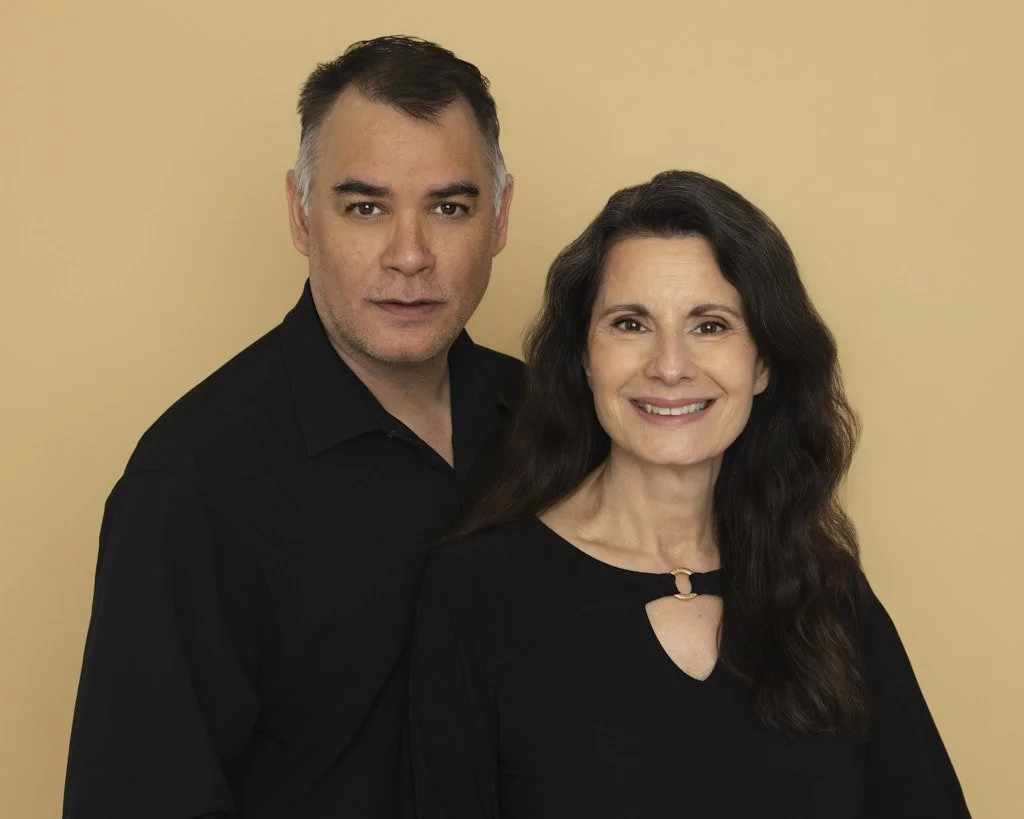 Portrait of Photographers Kendell and Evelyn Healy standing together against a light yellow background, both wearing black shirts. The man has short gray hair and the woman has long dark curly hair, and she is smiling.