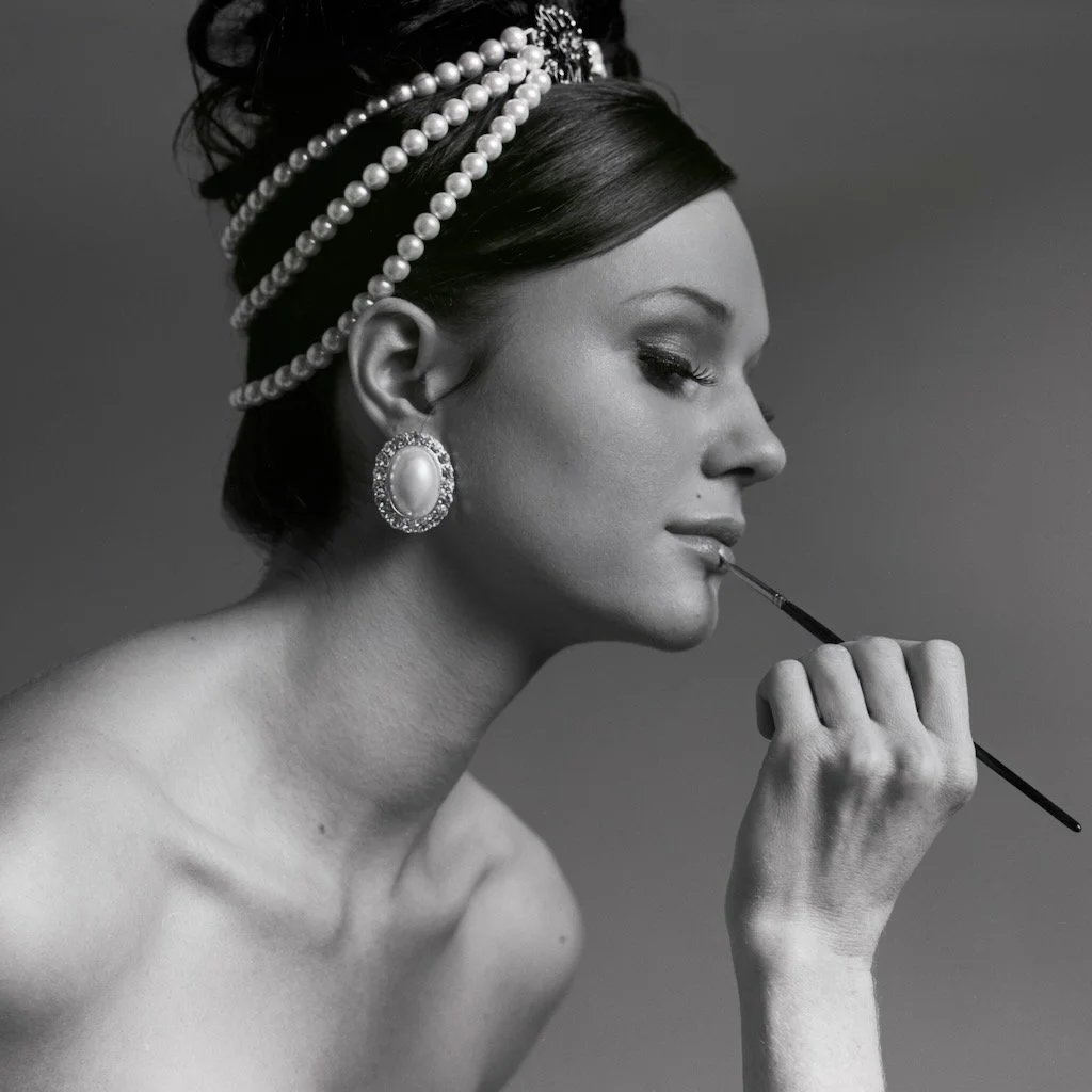 A woman with pearl jewelry and earrings, applying makeup with a brush in a black and white photo.