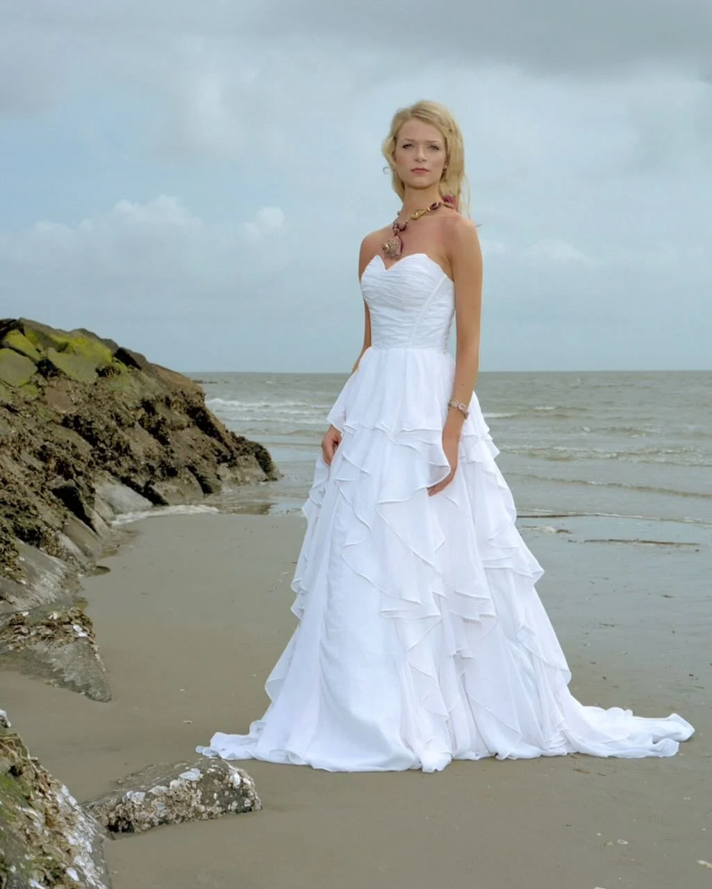 A woman in a white wedding dress standing on a beach with rocks and the ocean in the background.