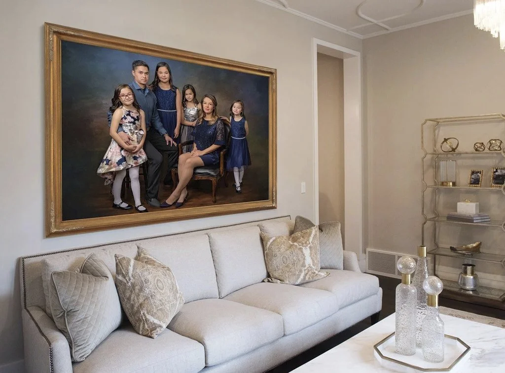 A large family portrait hangs above a cream-colored sofa in a well-decorated living room. The portrait features a man, woman, and five young girls, with the woman sitting on a chair and the rest standing around her.