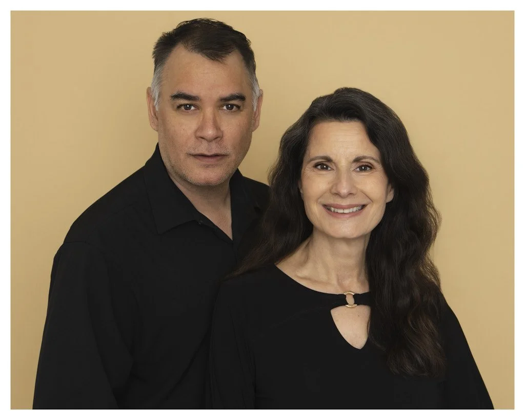 A middle-aged man and woman stand together against a beige background, both wearing black tops. The man has short, dark hair with some gray and serious expression; the woman has long, wavy dark hair and is smiling.