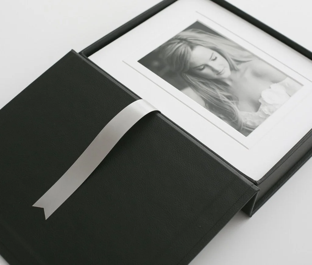 A black photo album with a white ribbon, containing a black and white portrait of a young woman with long hair.