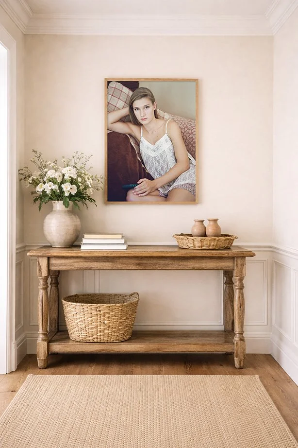 A wooden console table against a wall with a large framed portrait of a young woman in a white lace dress, leaning on a sofa. On the table are a large white vase with white flowers, a stack of white books, and a wicker basket with two small clay pots. There is a wicker basket on the lower shelf of the table and a beige rug on the floor.