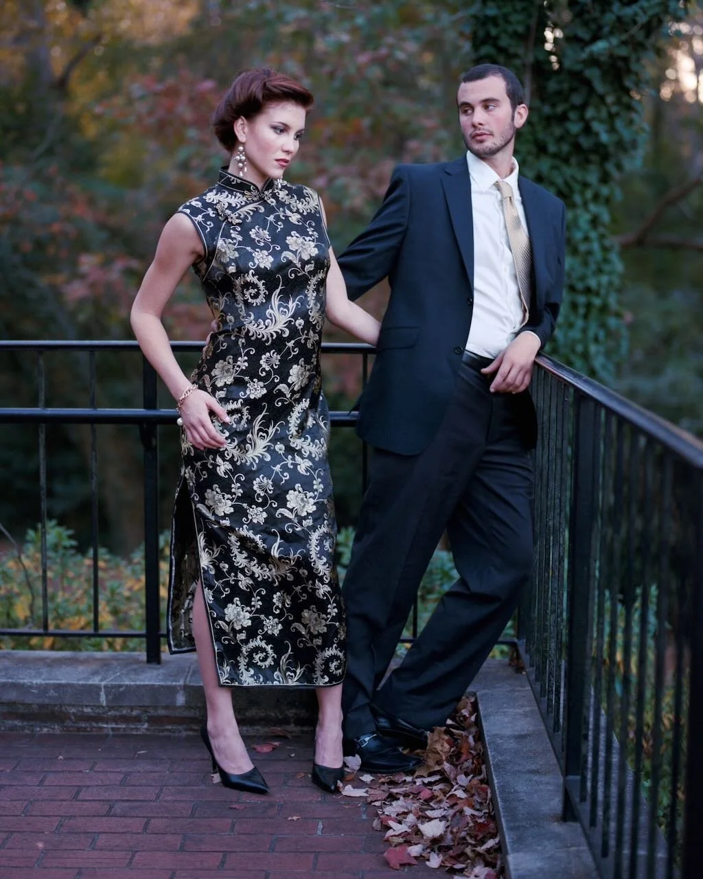 A man and woman dressed in formal attire standing on a balcony with a wooded background, autumn leaves on the ground.