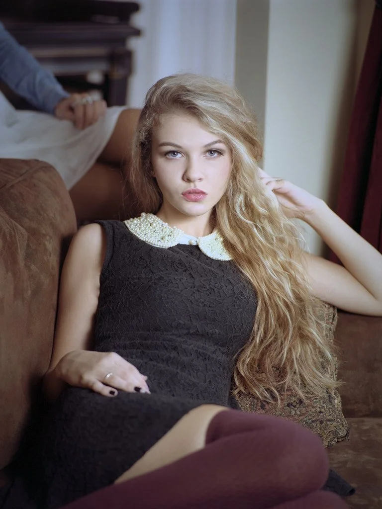 blonde young woman lounging wearing a black dress with a white collar and red stockings.jpg