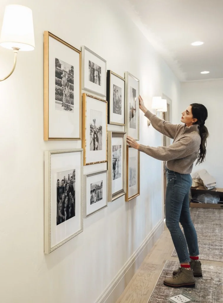 A woman hanging framed black-and-white photographs or artwork on a white wall in a well-lit room.