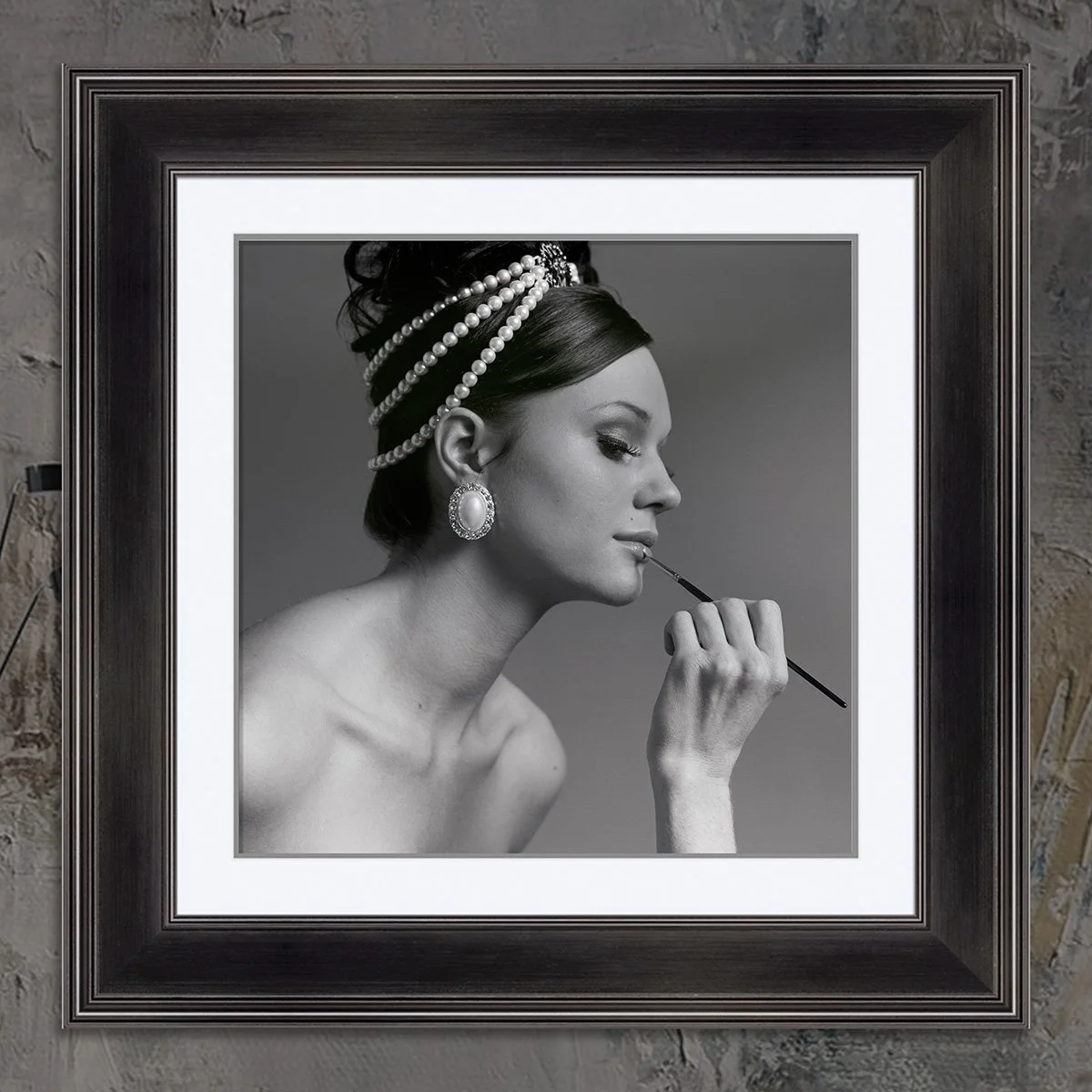 Black and white framed photograph of a woman with pearl jewelry, holding a makeup brush near her lips, with a hairstyle adorned with pearls, displayed on a brick wall.