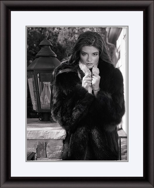 Black-and-white portrait of a woman with long hair, wearing a fur coat and holding the collar close to her face, standing outdoors near a stone wall and lantern.