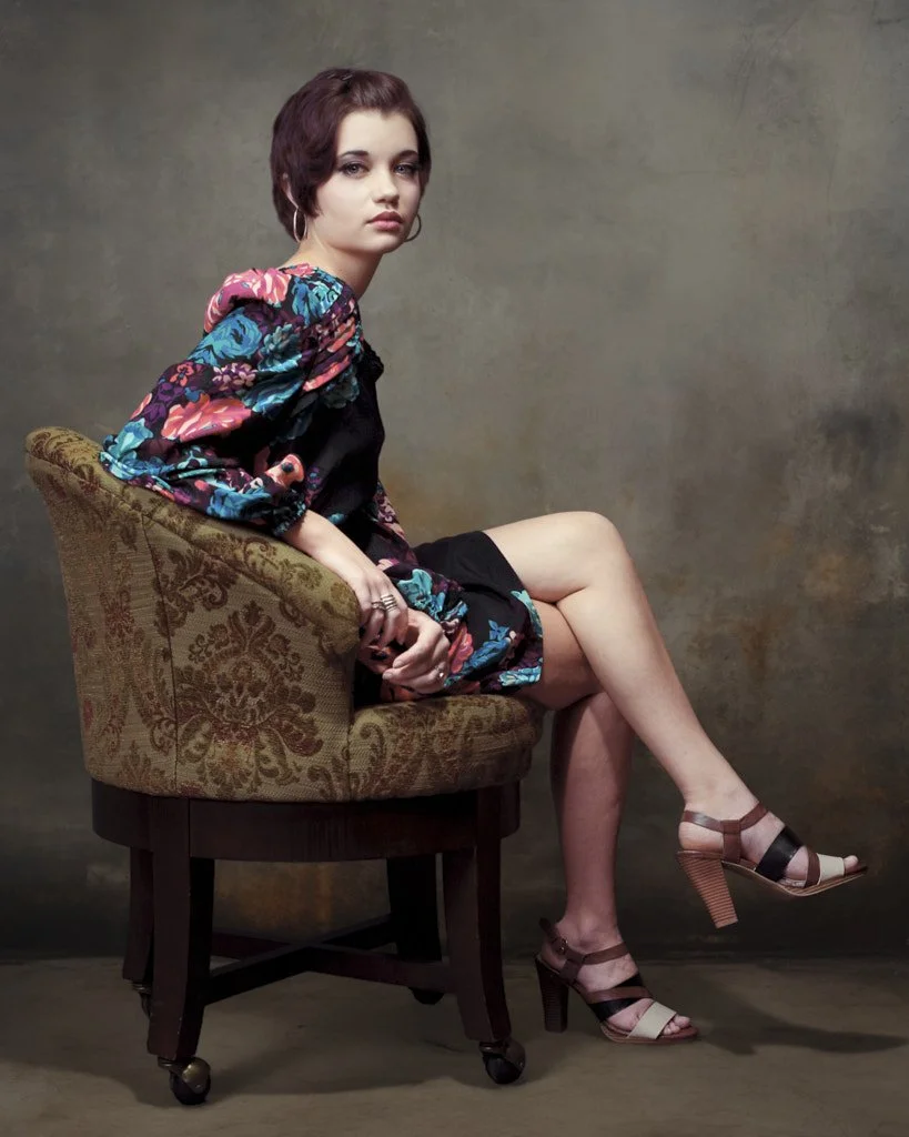 A young woman with short dark hair and hoop earrings sitting sideways in a patterned armchair, wearing a colorful floral jacket, black dress, and high heeled sandals against a neutral background.