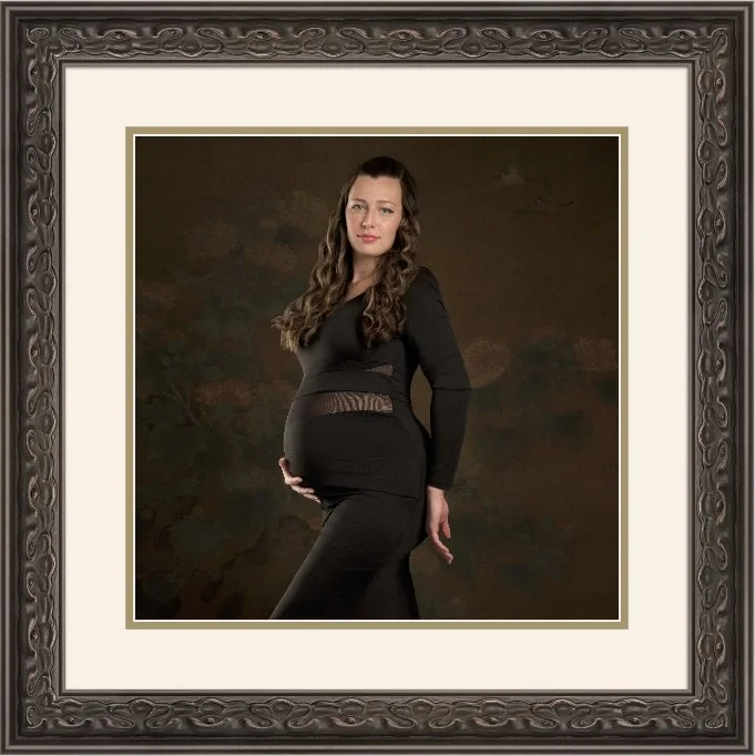 A pregnant woman with long wavy hair, wearing a form-fitting black dress, poses against a dark background in a decorative framed studio portrait.
