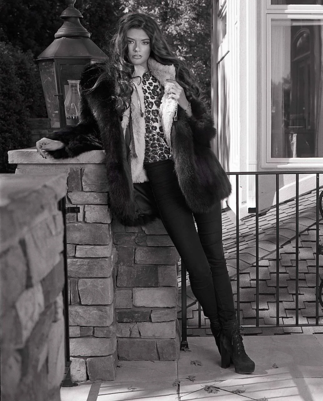 A woman with long wavy hair wearing a leopard print top, leather pants, a fur coat, and high-heeled boots leaning against a stone pillar on an outdoor porch.