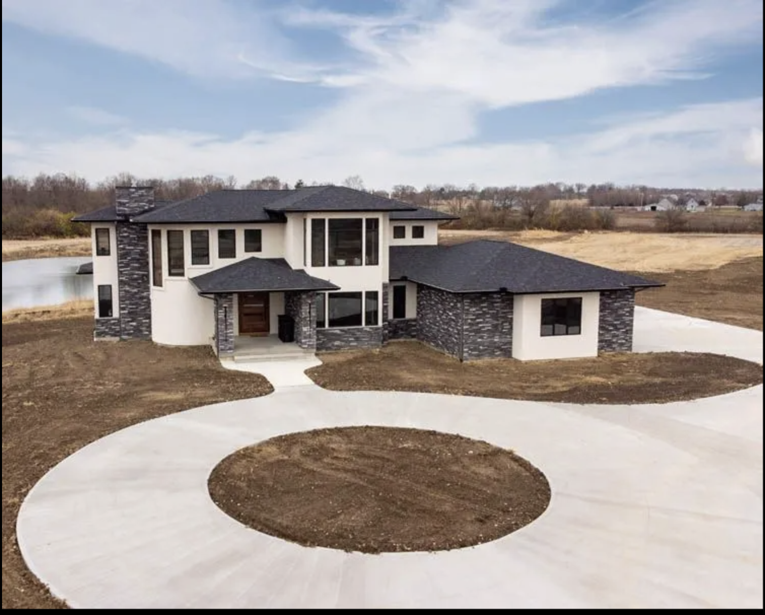 A modern two-story house with a black shingle roof, dark stone accents on the exterior, large windows, and a circular concrete driveway leading to the front entrance, set in a rural area with a pond and open land.