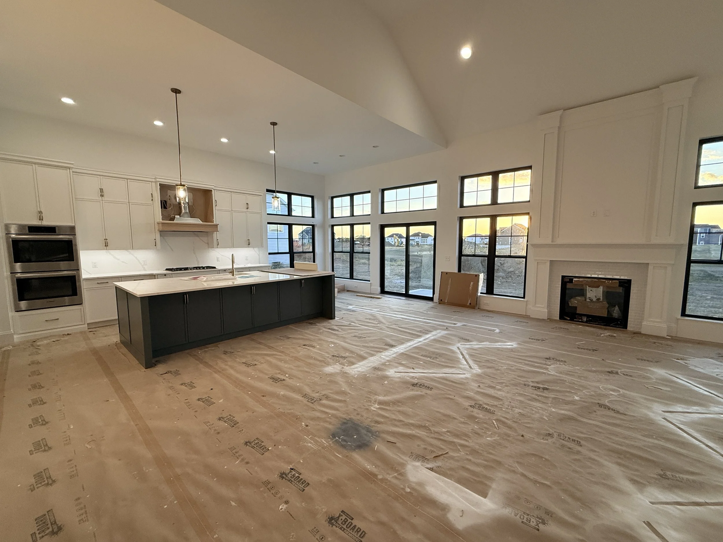 Open-concept kitchen and living room under construction with large windows, island, and fireplace.