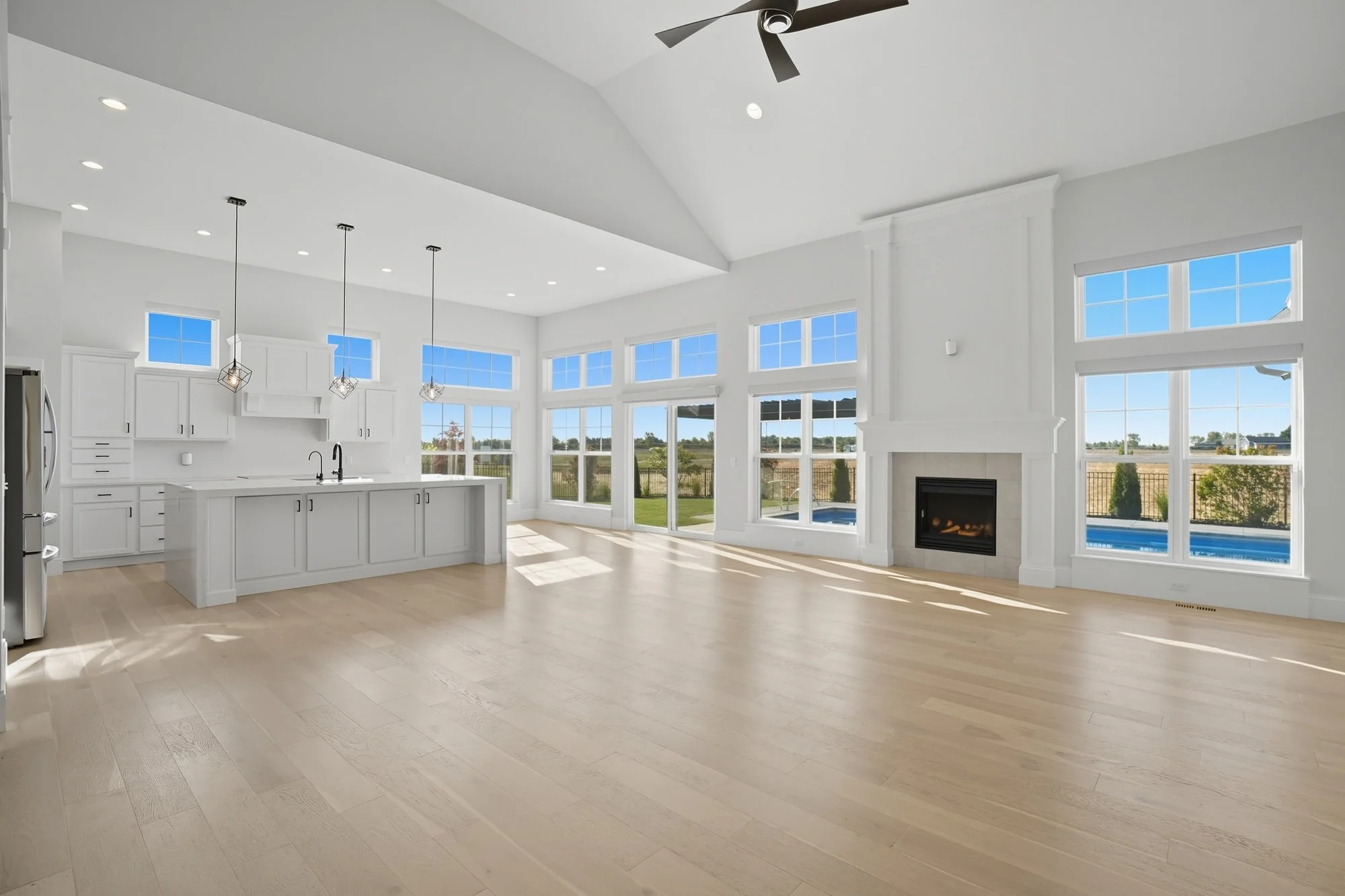 Empty modern kitchen and living room with large windows, white cabinets, a fireplace, and light wood flooring.