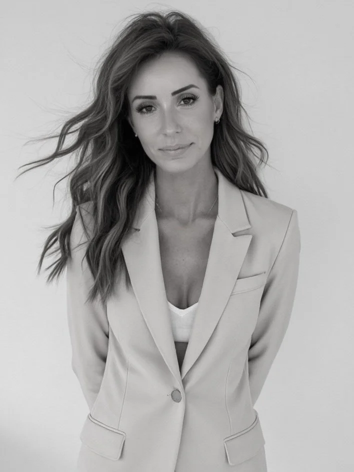 Black and white portrait of a woman with wavy hair, wearing a blazer over a top, standing against a plain background.