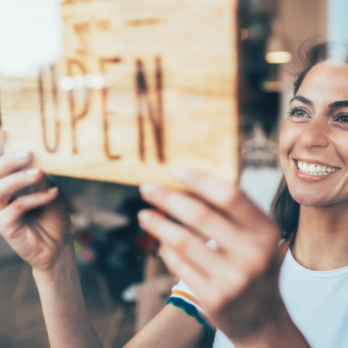 Woman smiling and hanging a wooden “We’re Open” sign.