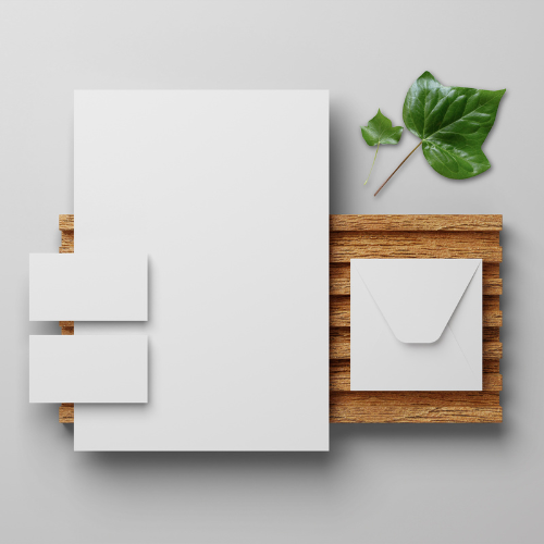 Flat lay of a wooden tray with blank white paper, envelopes, and sticky notes, accompanied by green leaves on a gray background.