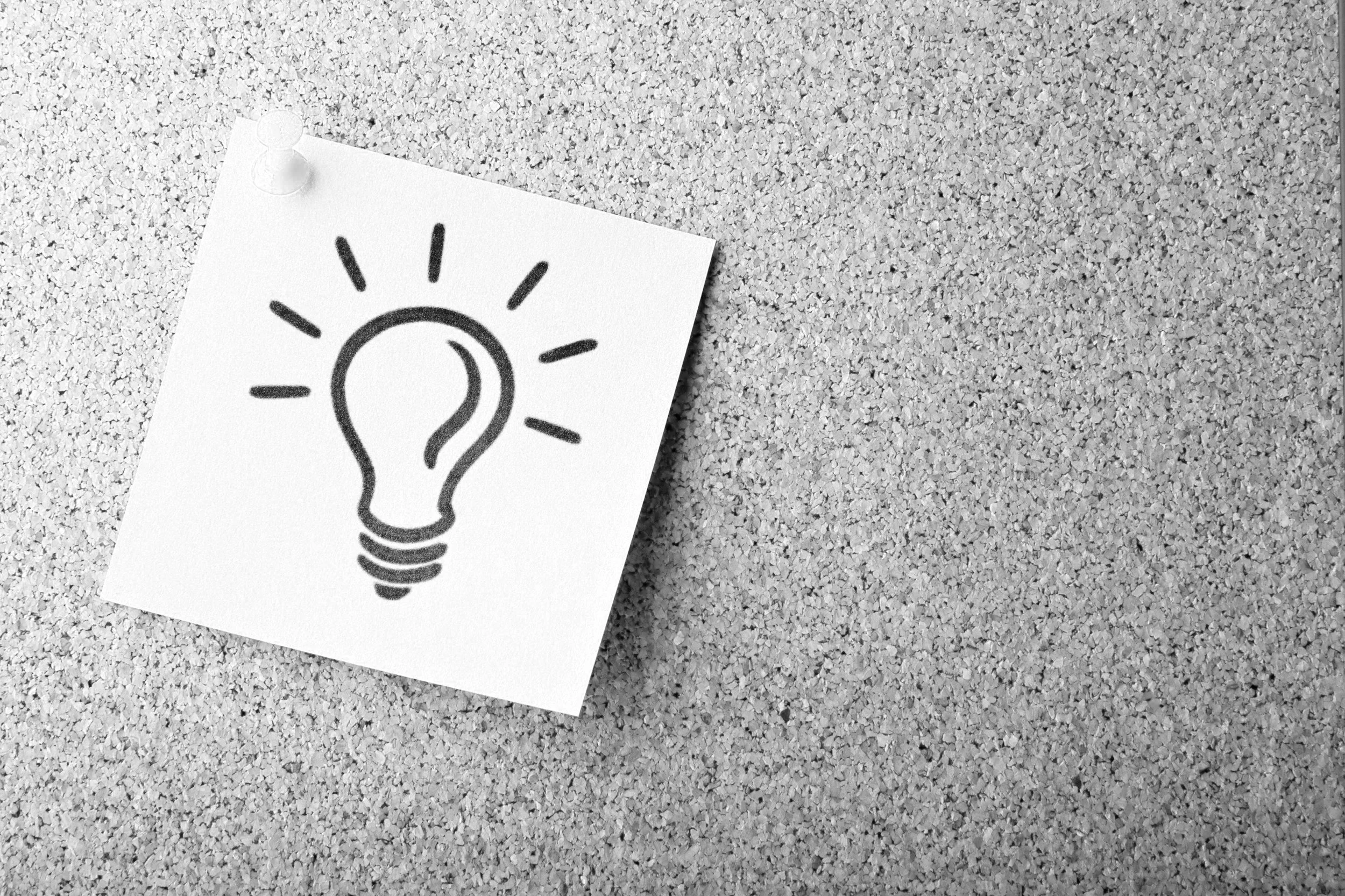A white note pinned to a corkboard with a drawing of a light bulb and lines indicating illumination.