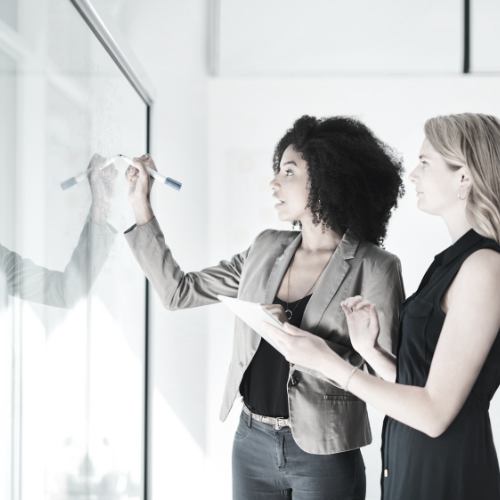 Two women in business attire writing on a glassboard with a marker in an office setting.