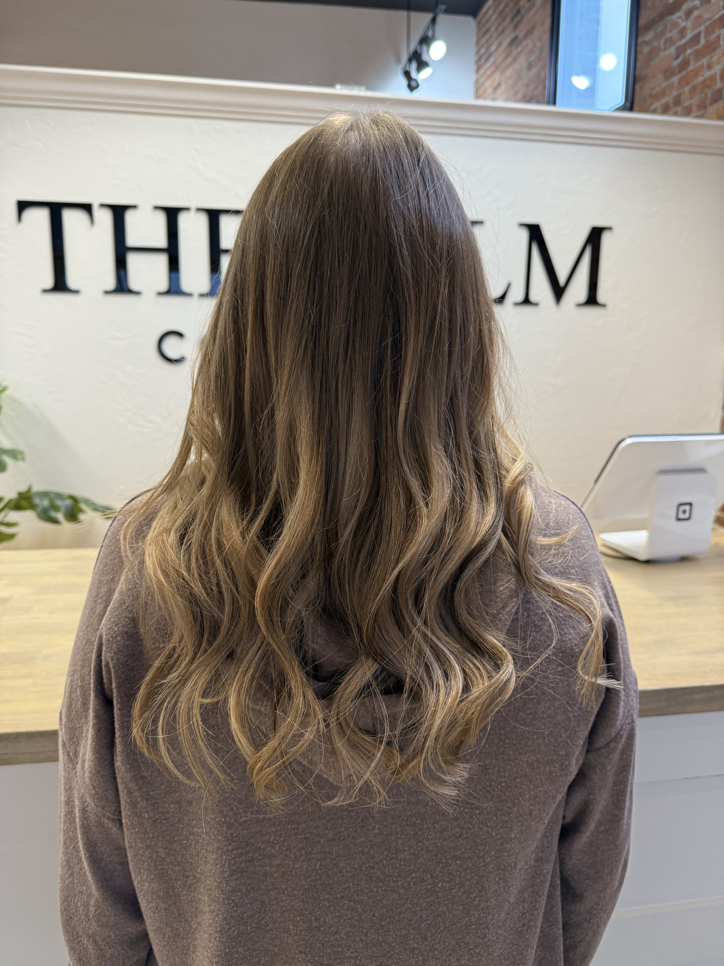 Back of a woman with long, wavy, light brown hair sitting at a wooden counter in a salon, with a white wall and black text in the background.