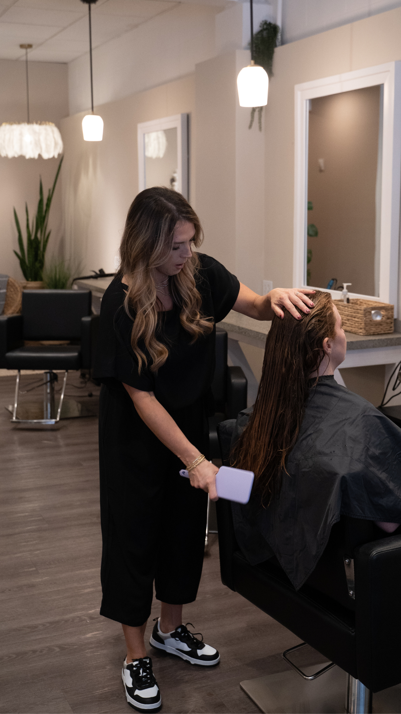 A hairstylist applies hair treatment to a client's wet hair in a salon with modern decor, hanging pendant lights, and large mirrors.