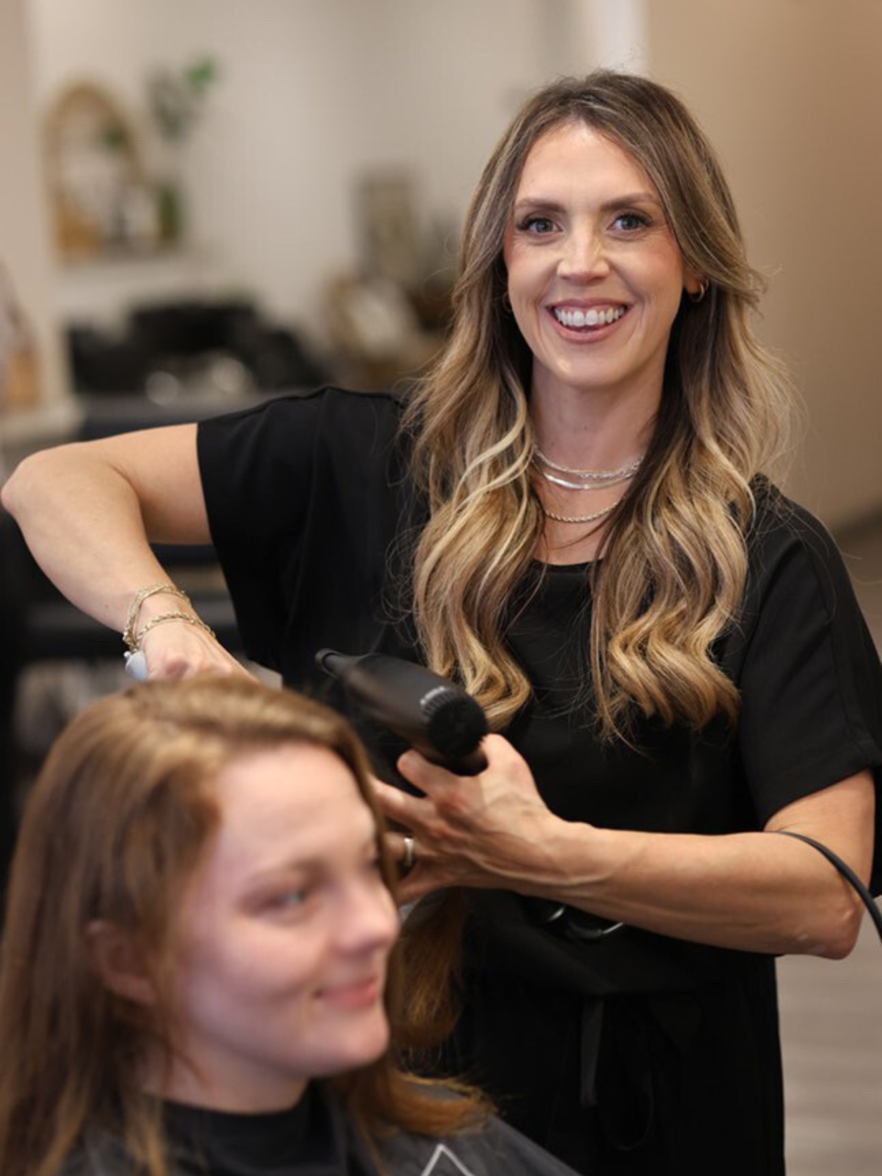 A woman with long, wavy hair smiling while drying a young woman's hair at a salon.