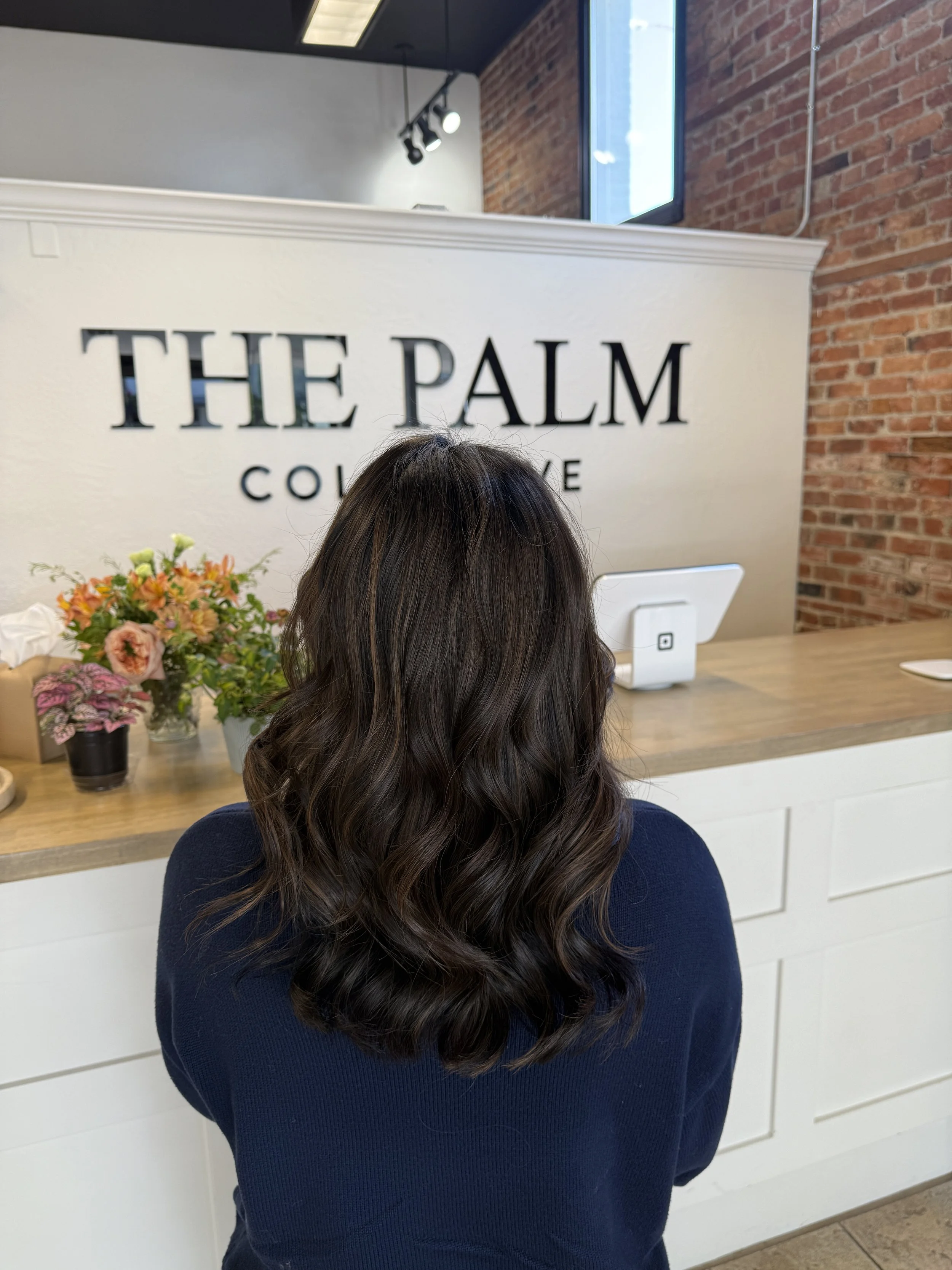 Back of a woman with wavy brown hair standing at a counter in a salon called The Palm Coffee, with a brick wall and a sign visible in the background.