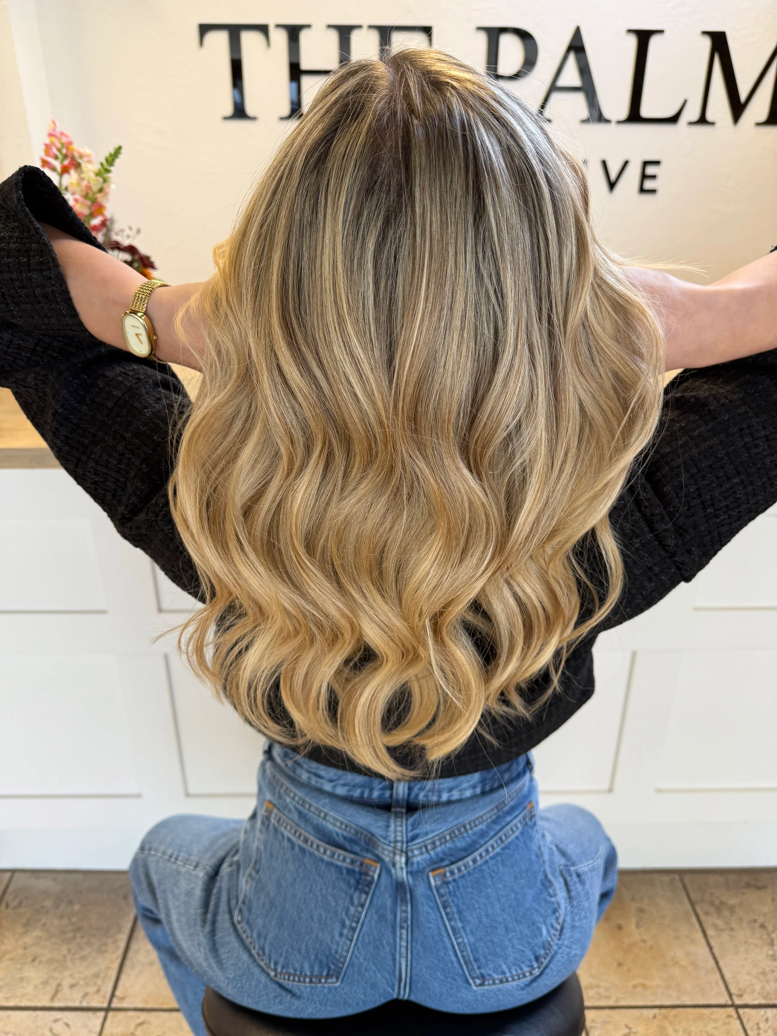 Back view of a woman with long, wavy blonde hair sitting on a stool in a salon with white cabinets and a floral arrangement in the background.