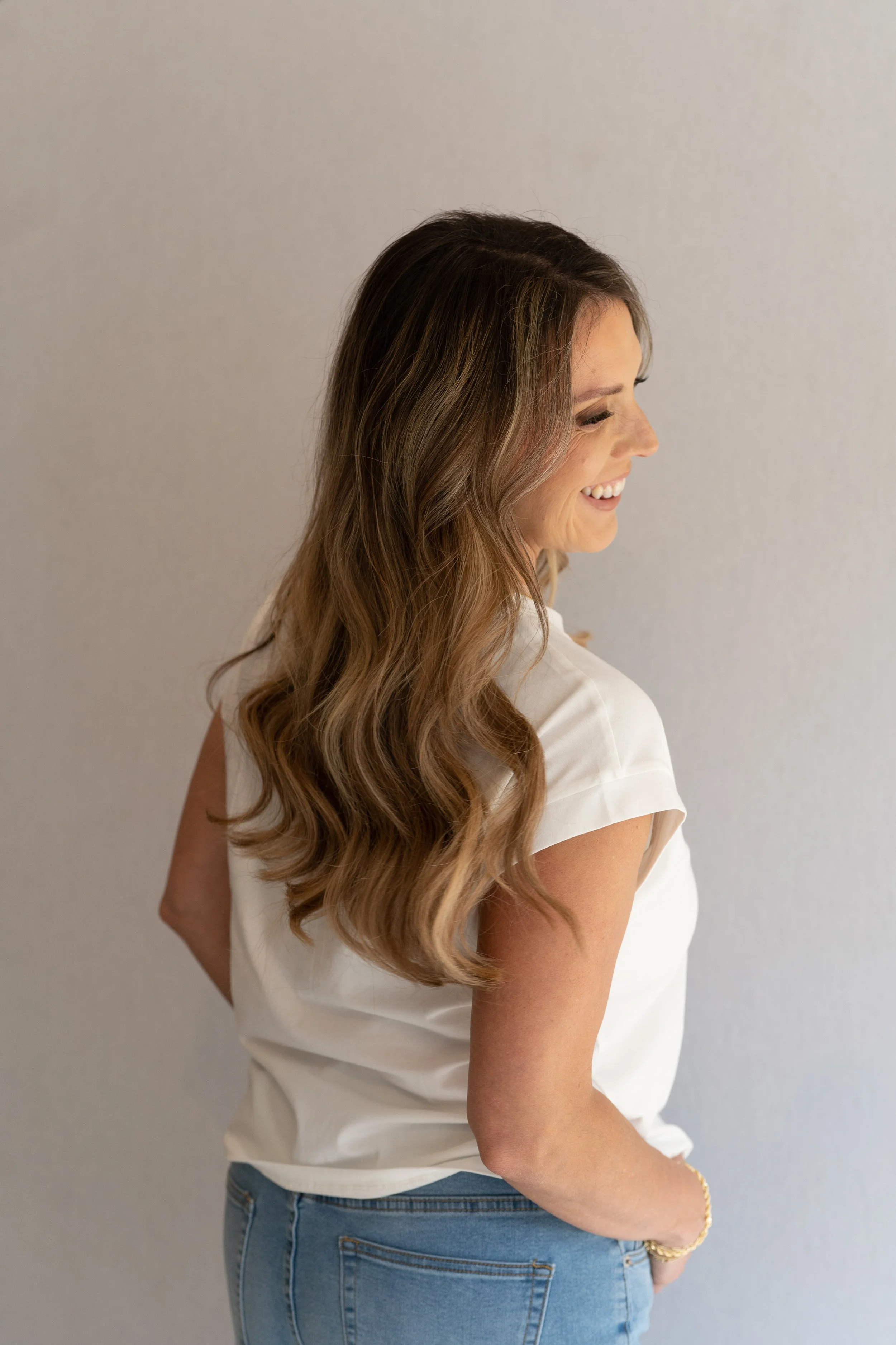 Smiling woman with long wavy hair wearing a white top and blue jeans, standing sideways against a plain wall
