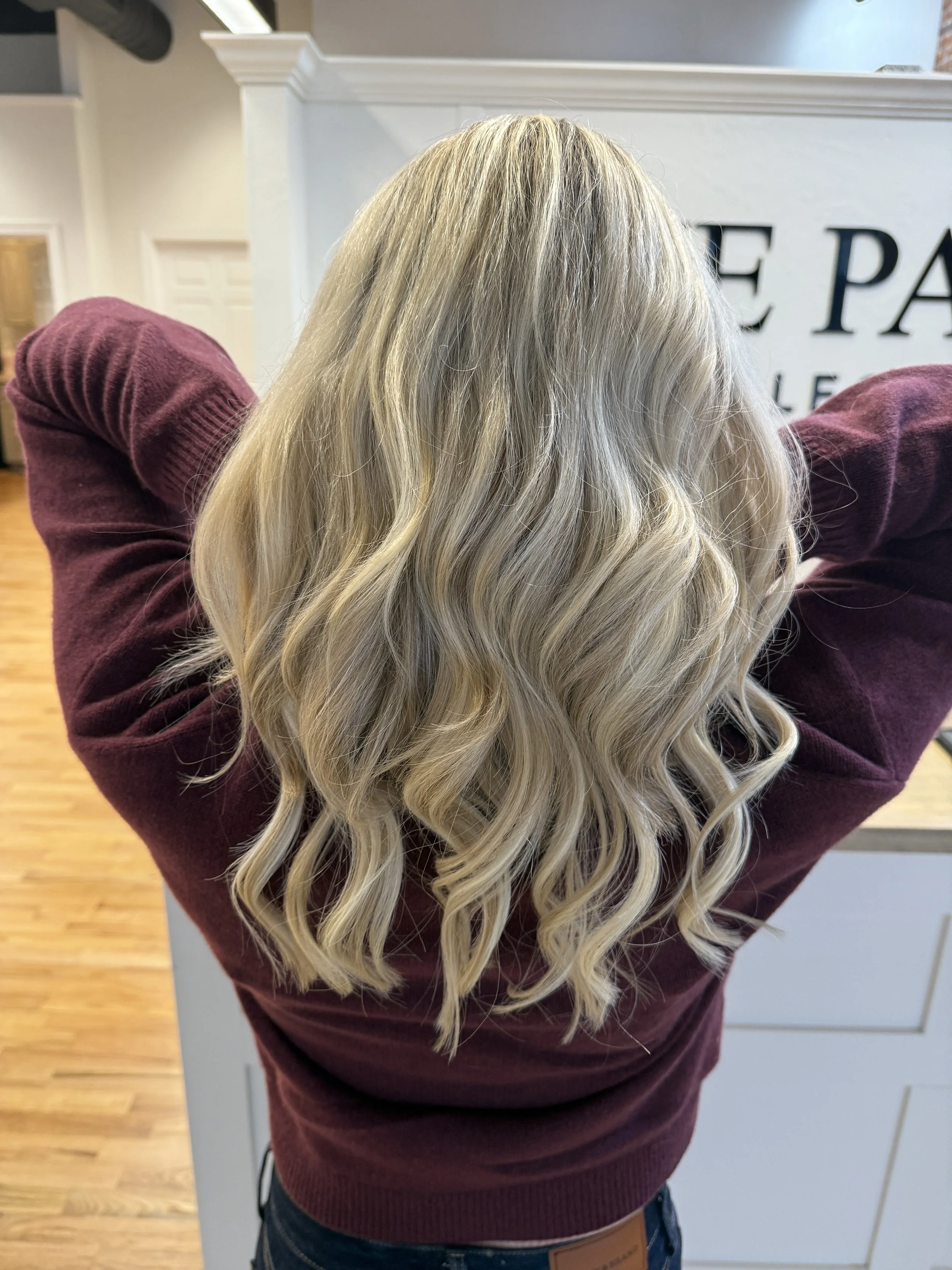 Back of a woman with long, wavy, blonde hair wearing a maroon sweater. She is standing in a room with a white wall and a white doorway in the background.