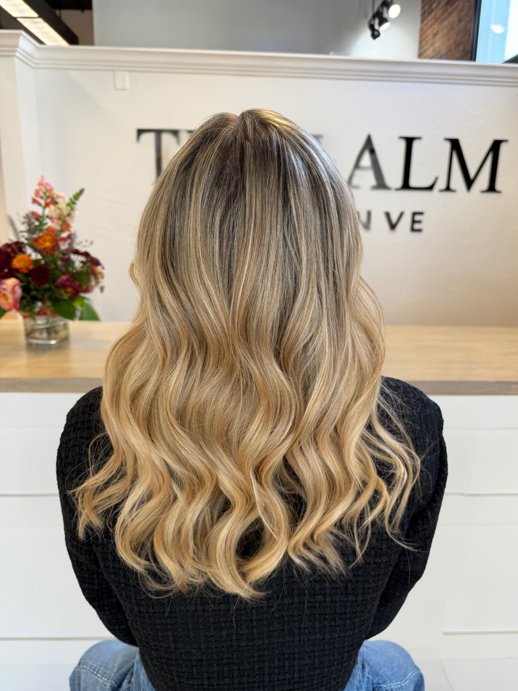 Back view of a woman with long, wavy blonde hair sitting in a salon.