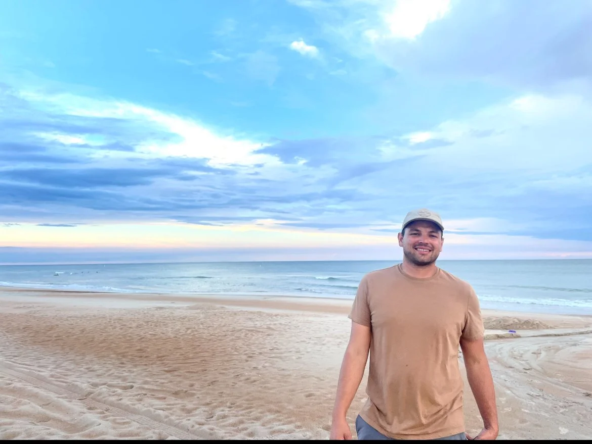 Smiling man standing on a sandy beach with ocean waves and a cloudy sky in the background.