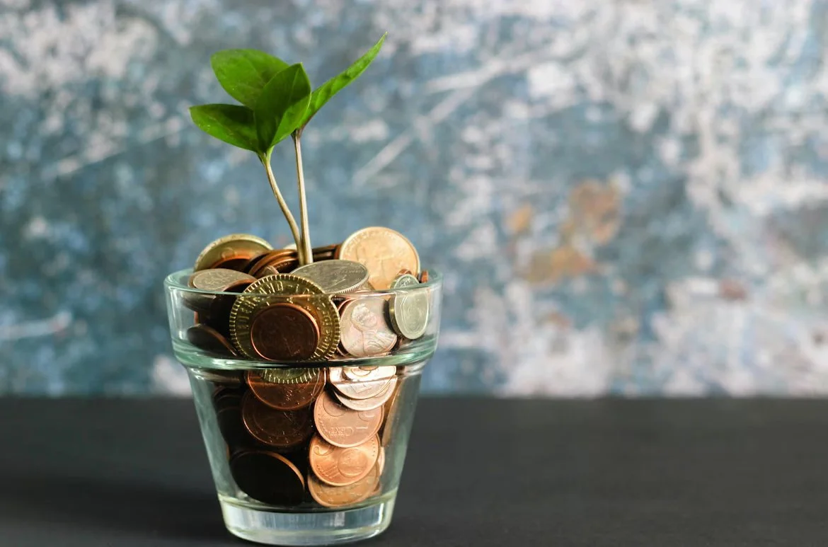 A glass filled with coins of various denominations and a small green plant growing out of the coins.
