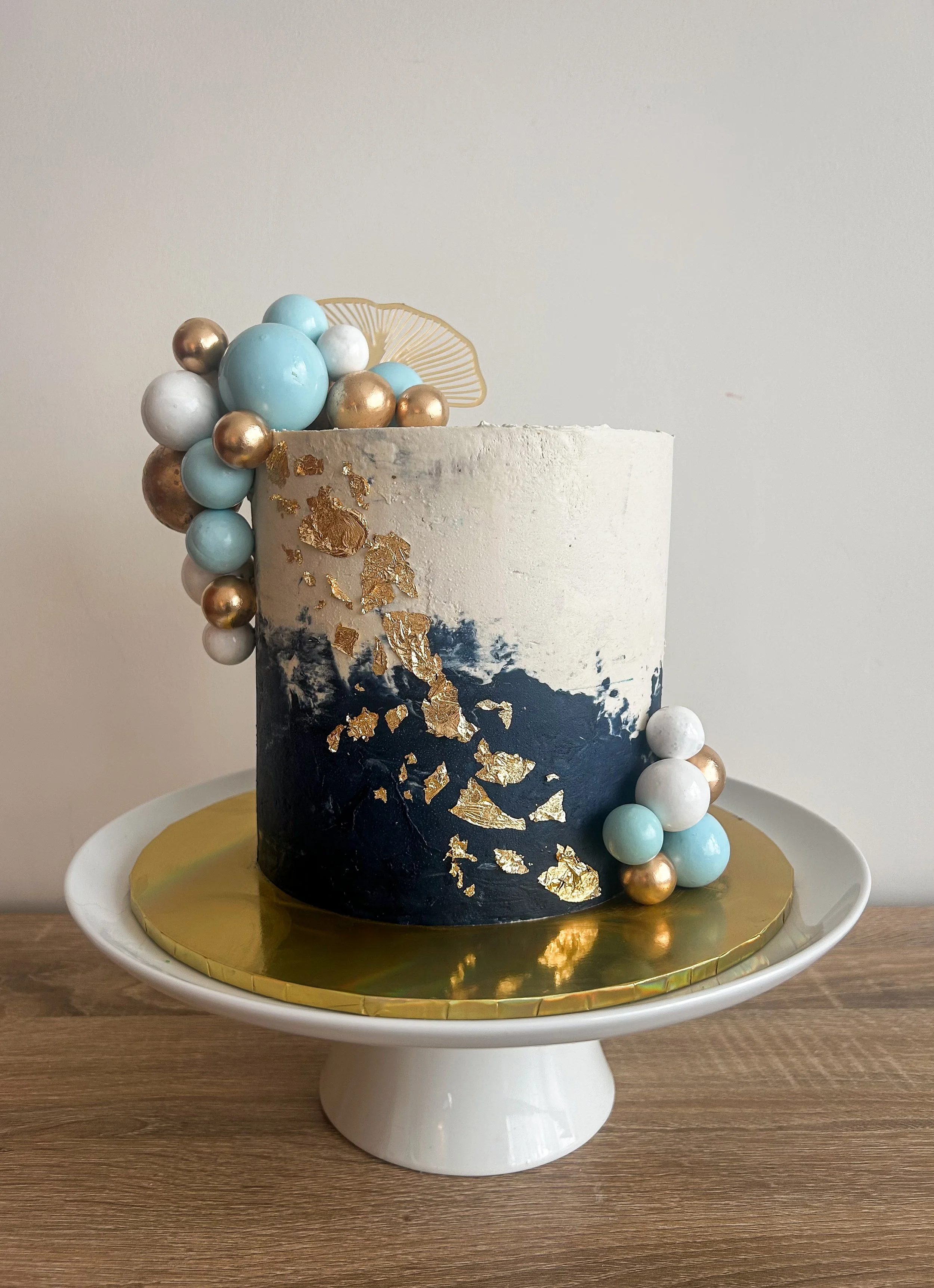 A round, tall cake with a black and white abstract design, decorated with gold leaf accents, on a white cake stand with a gold-colored base. The cake is adorned with clusters of pastel blue, white, and metallic gold spherical decorations on top and a