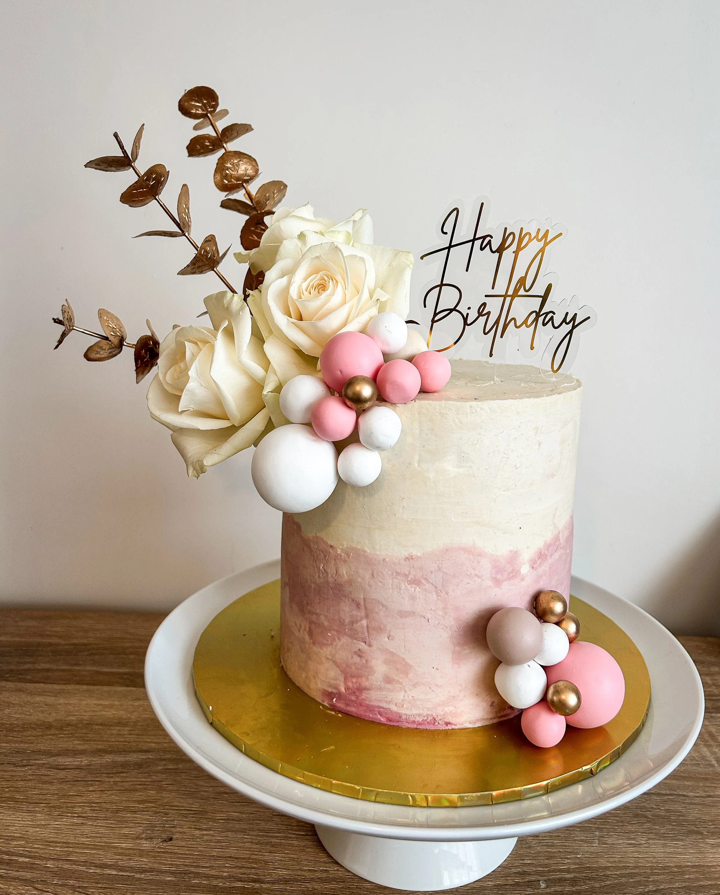 A birthday cake decorated with white, pink, and gold spheres, white roses, and a "Happy Birthday" topper on a white cake stand.