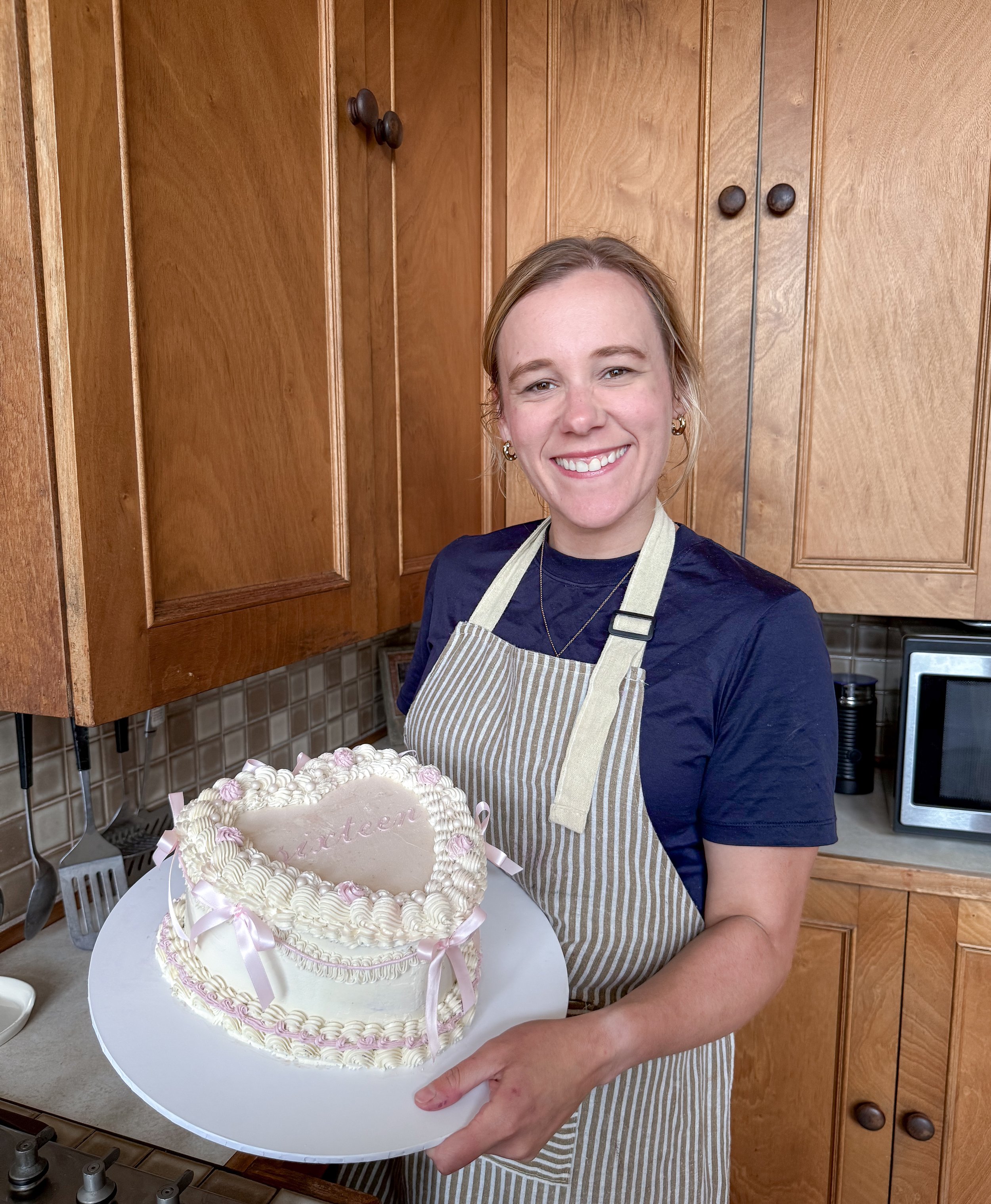 Woman smiling and holding a heart-shaped cake decorated with white frosting and pink ribbons in a kitchen.
