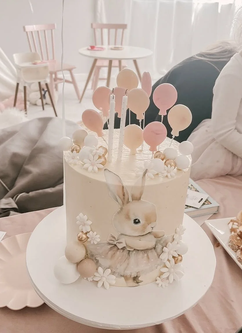 A pink and white birthday cake decorated with a bunny illustration, surrounded by flowers, balloons, and candles. The cake is on a white cake stand, with a pink table, chairs, and a person in the background.